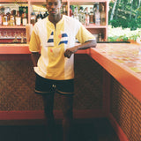 A man wearing a yellow and white short-sleeve athletic jersey with blue and red accents, paired with black athletic shorts, standing in a tropical bar environment.
