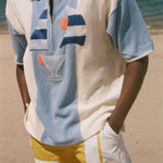 A man wearing a light blue and off-white color-blocked short-sleeve polo shirt with a half-zip collar, featuring a graphic print of a sailboat and a cocktail glass on the front. He is also wearing yellow and white shorts, standing on a sandy beach with the ocean in the background.