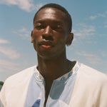 A man wearing a white zip-up shirt with blue and orange graphic details, photographed outdoors against a blue sky with light clouds.
