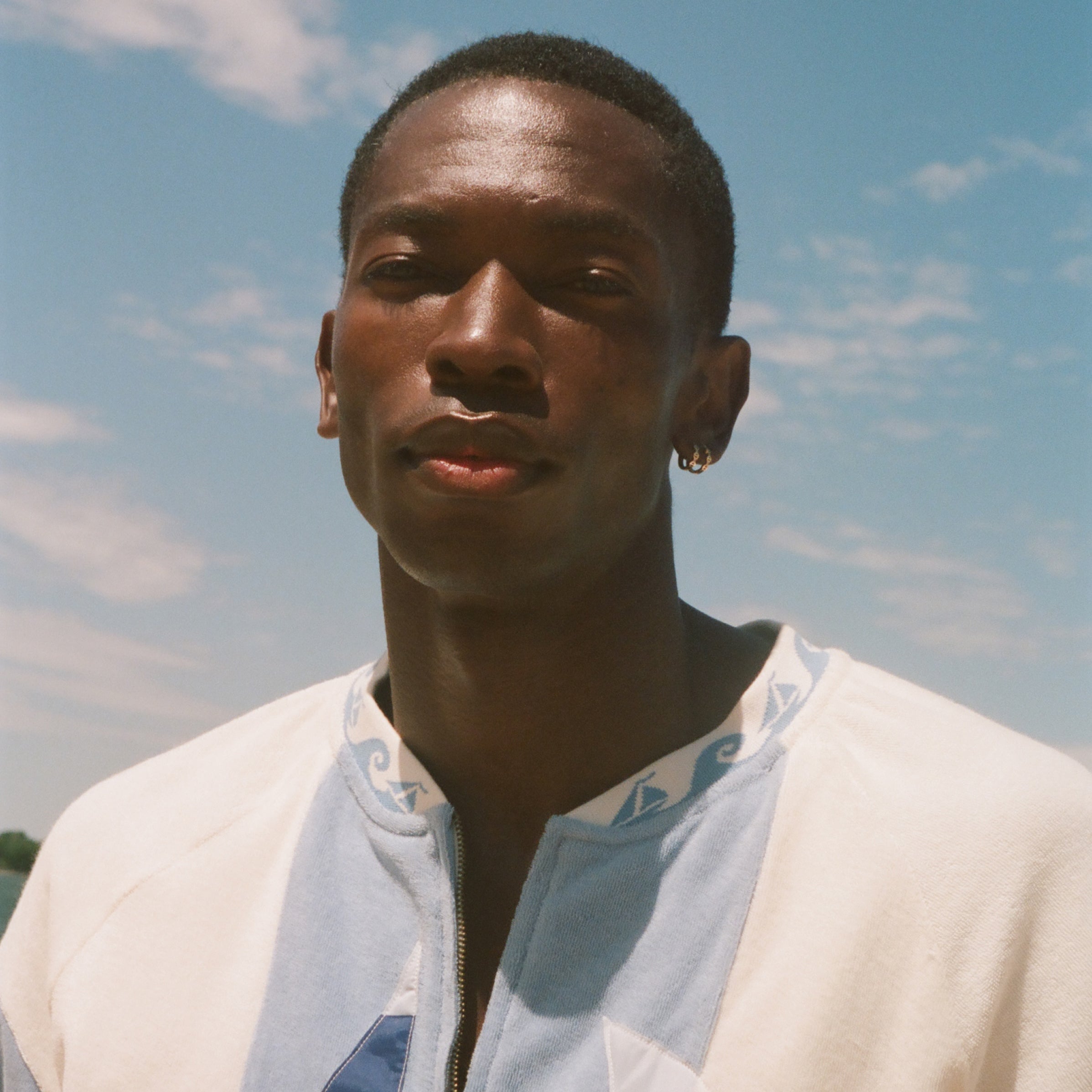 A man wearing a white zip-up shirt with blue and orange graphic details, photographed outdoors against a blue sky with light clouds.