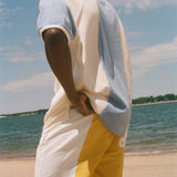 A man standing on a sandy beach wearing a color-blocked short-sleeve cabana shirt. The shirt features cream, light blue, and gray color blocking, while the shorts are white with a yellow side panel.