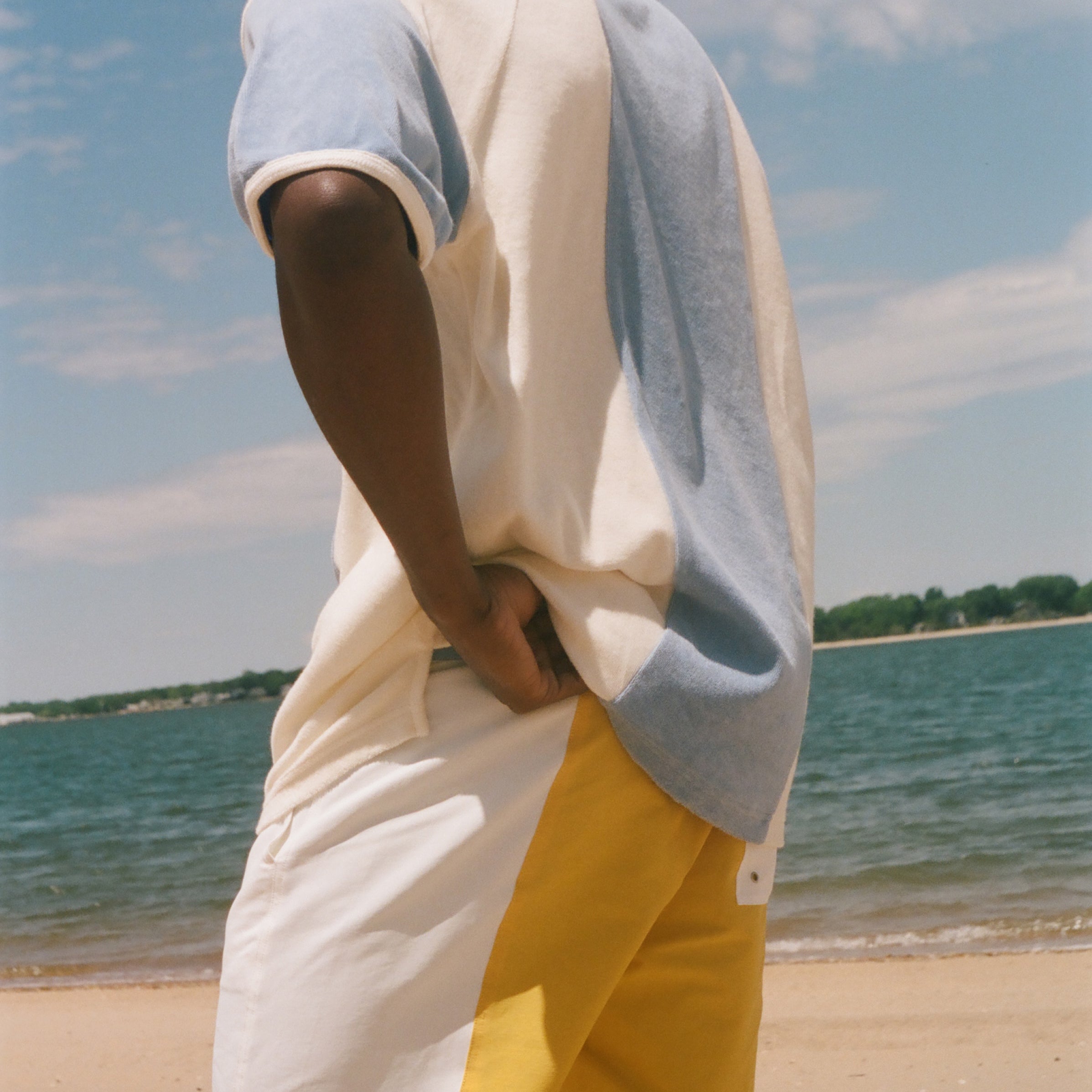 A man standing on a sandy beach wearing a color-blocked short-sleeve cabana shirt. The shirt features cream, light blue, and gray color blocking, while the shorts are white with a yellow side panel.