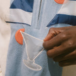 A close-up of a person wearing a light blue zip-up jacket with navy and white stripes, orange circular patches, and a white embroidered cocktail glass motif, while holding a small white fabric piece.