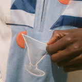 A close-up of a person wearing a light blue zip-up jacket with navy and white stripes, orange circular patches, and a white embroidered cocktail glass motif, while holding a small white fabric piece.