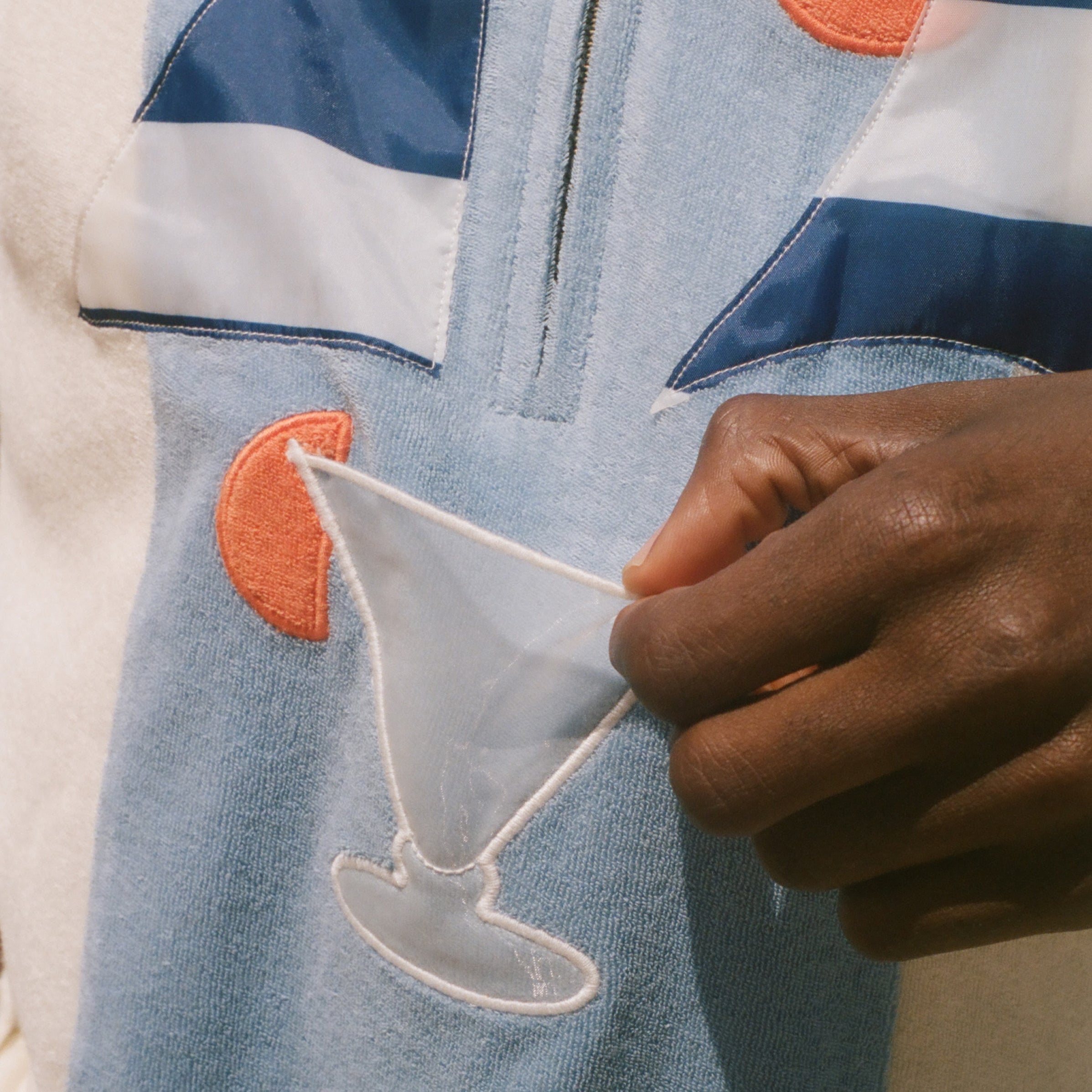 A close-up of a person wearing a light blue zip-up jacket with navy and white stripes, orange circular patches, and a white embroidered cocktail glass motif, while holding a small white fabric piece.