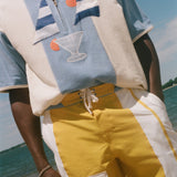 A person standing on a beach wearing a light blue and cream short-sleeve polo shirt with embroidered sailboats and a martini glass, paired with yellow and white drawstring shorts with a white cargo pocket.