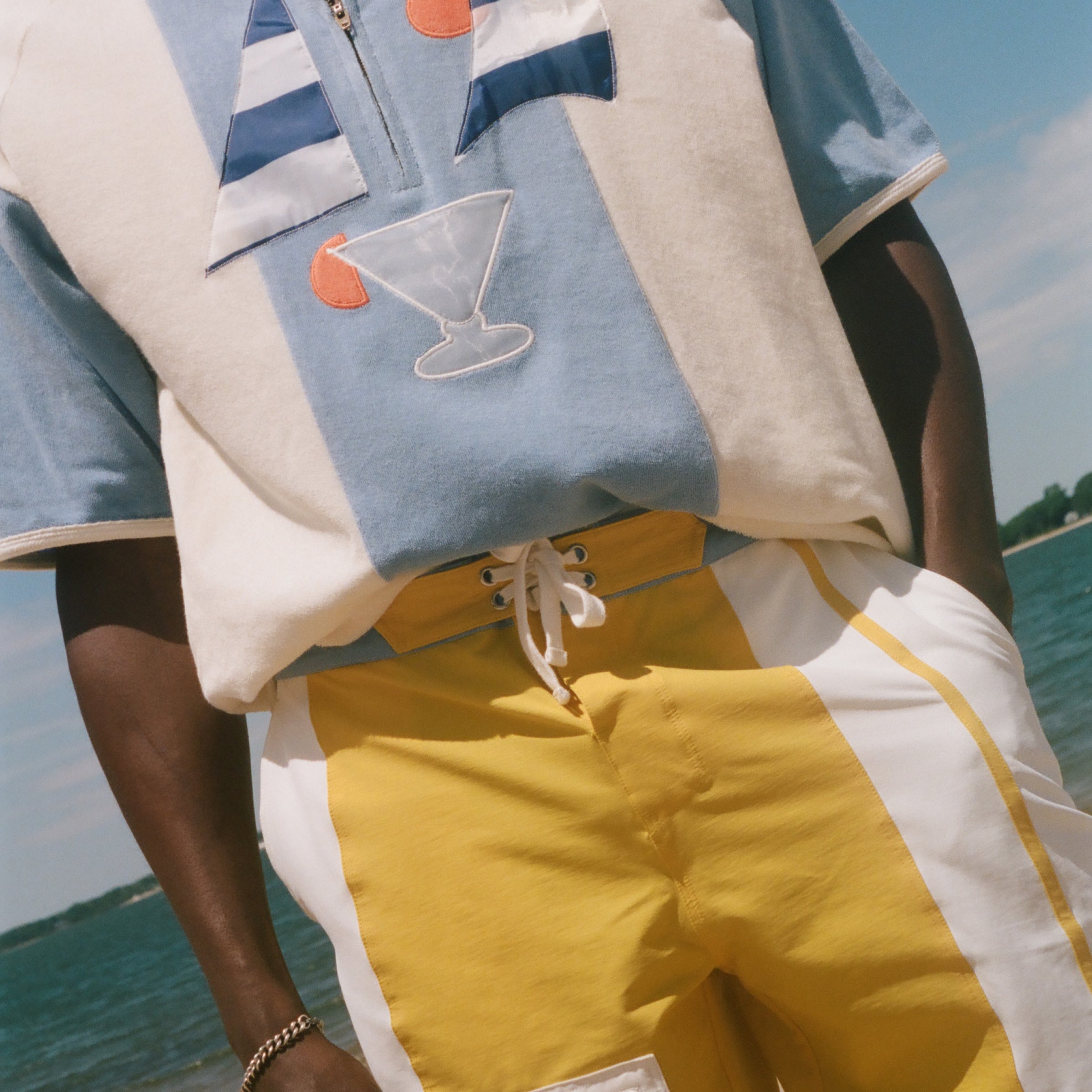 A person standing on a beach wearing a light blue and cream short-sleeve polo shirt with embroidered sailboats and a martini glass, paired with yellow and white drawstring shorts with a white cargo pocket.