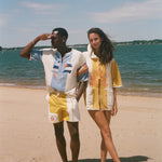 A man and a woman standing on a sandy beach wearing matching vintage-style swimwear sets. The man wears a white and light blue short-sleeve collared swim shirt with yellow and white shorts, while the woman wears a yellow and white patterned short-sleeve cover-up shirt with matching yellow and white bottoms.