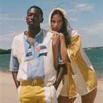 A man and a woman standing on a sandy beach wearing matching light blue and yellow resort wear, with the ocean and blue sky in the background.