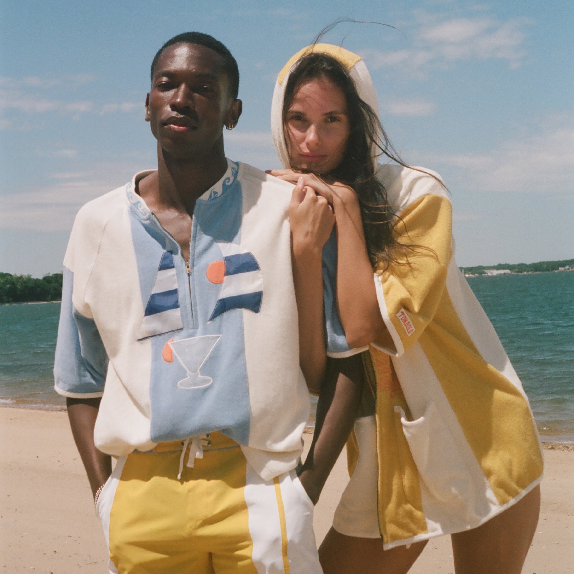 A man and a woman standing on a sandy beach wearing matching light blue and yellow resort wear, with the ocean and blue sky in the background.