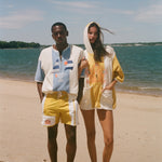 Two models standing on a sandy beach wearing matching yellow and off-white hooded beach poncho-style tops under a blue sky.
