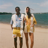 Two models standing on a sandy beach wearing matching yellow and off-white hooded beach poncho-style tops under a blue sky.
