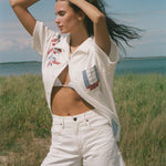 Person by the water wearing a cream terry cabana shirt with embroidered floral and window motifs, styled with white shorts.