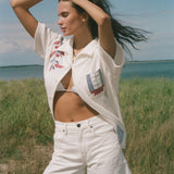 Person by the water wearing a cream terry cabana shirt with embroidered floral and window motifs, styled with white shorts.