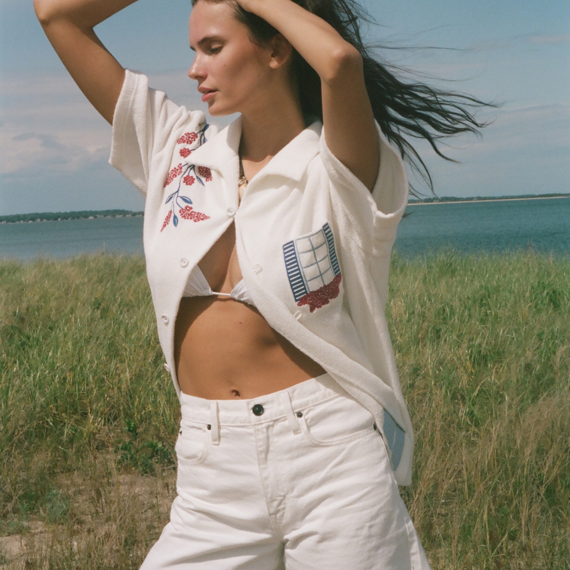 Person by the water wearing a cream terry cabana shirt with embroidered floral and window motifs, styled with white shorts.