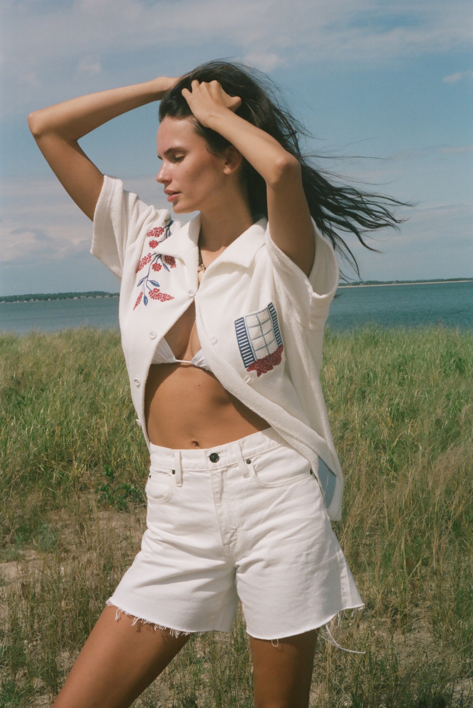 Person by the water wearing a cream terry cabana shirt with embroidered floral and window motifs, styled with white shorts.