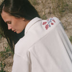 Back view of a person wearing a cream terry cabana shirt with embroidered red floral vines and blue leaves on the shoulder.
