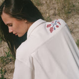 Back view of a person wearing a cream terry cabana shirt with embroidered red floral vines and blue leaves on the shoulder.