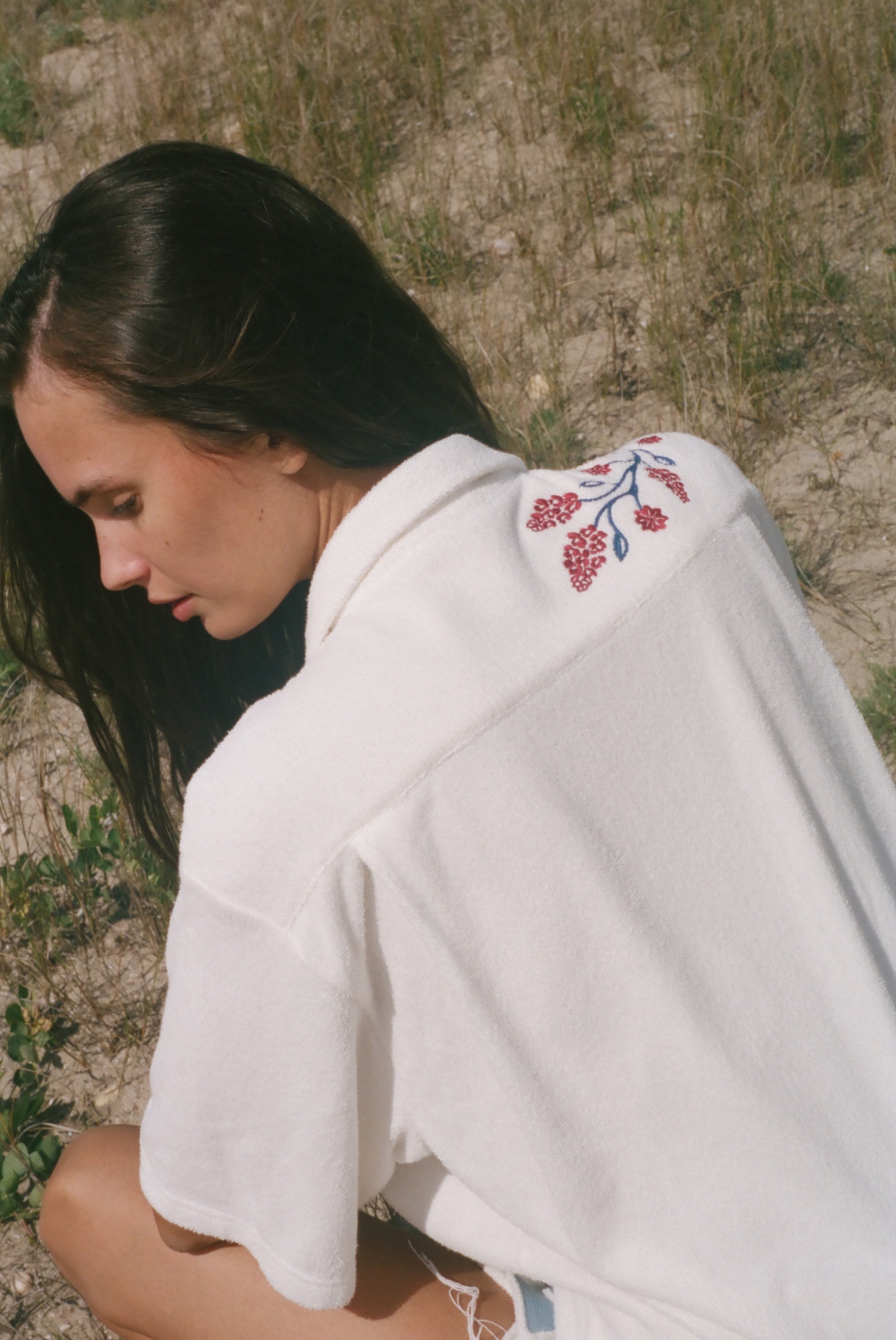 Back view of a person wearing a cream terry cabana shirt with embroidered red floral vines and blue leaves on the shoulder.