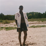 Person on a sandy beach wearing an open cream terry cabana shirt with embroidered floral, window, café, and cobblestone motifs.