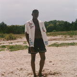 Person on a sandy beach wearing an open cream terry cabana shirt with embroidered floral, window, café, and cobblestone motifs.