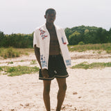 Man on a sandy beach wearing an open cream terry cabana shirt with embroidered floral, window, café, and cobblestone motifs.