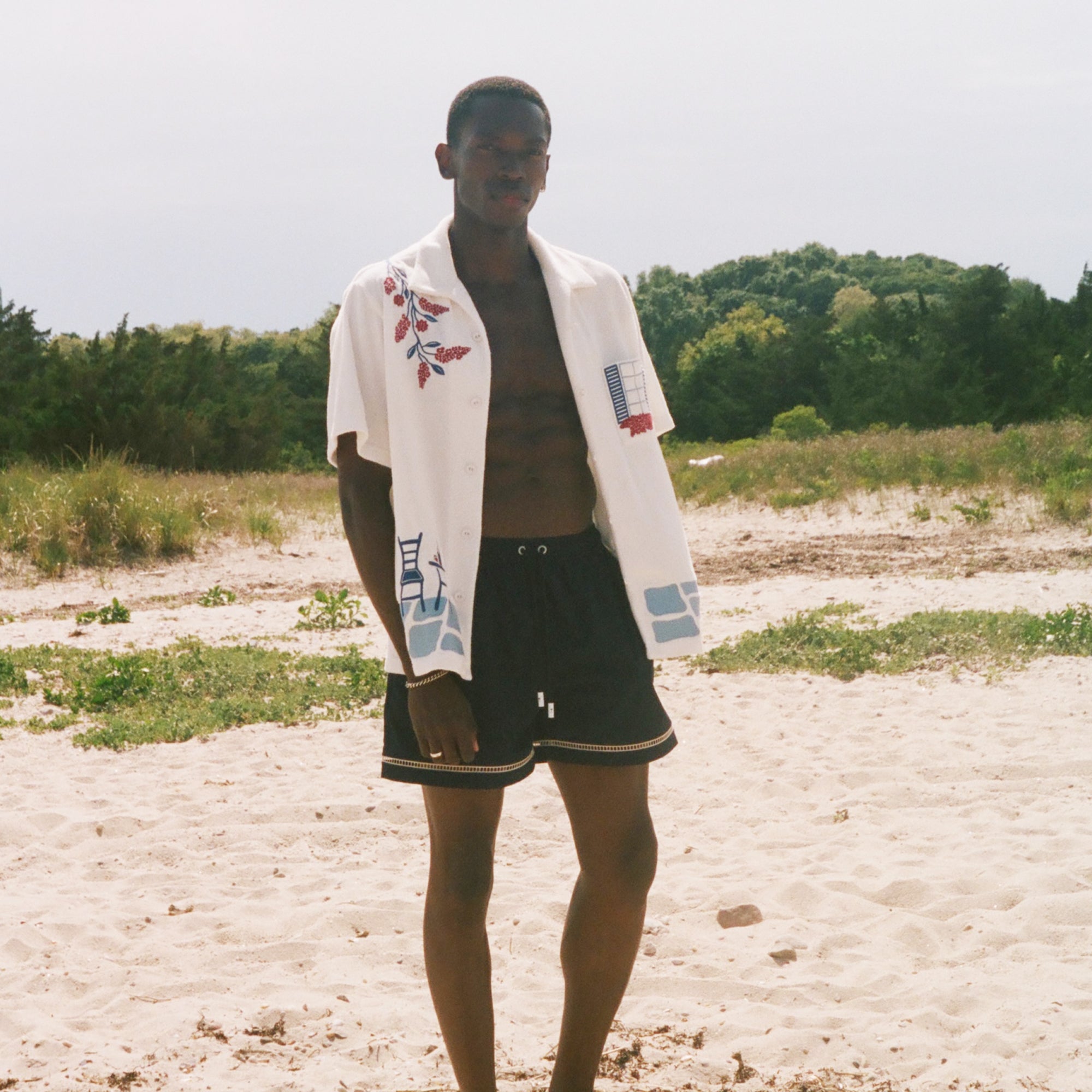 Man on a sandy beach wearing an open cream terry cabana shirt with embroidered floral, window, café, and cobblestone motifs.