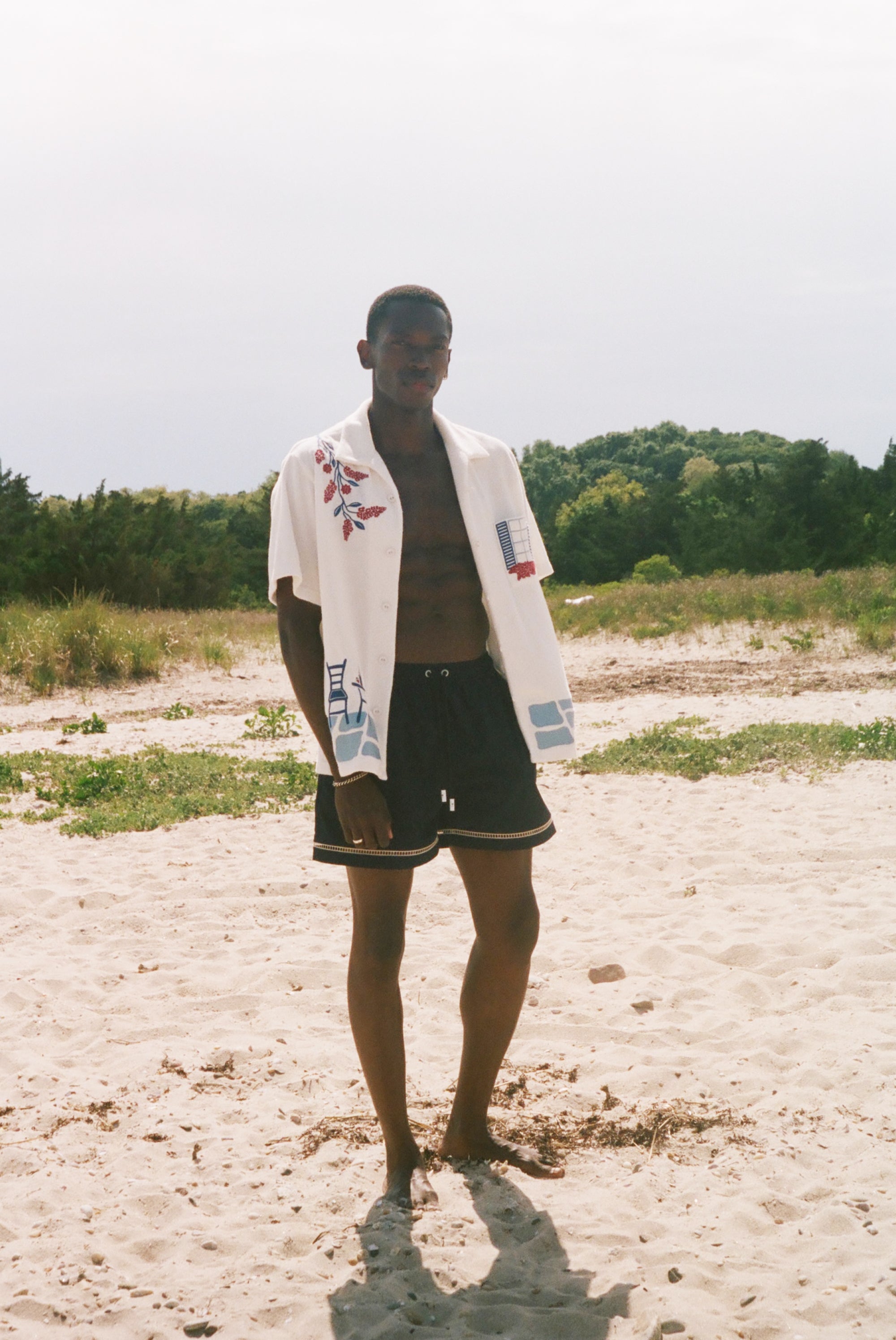 Man on a sandy beach wearing an open cream terry cabana shirt with embroidered floral, window, café, and cobblestone motifs.