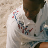 Overhead view of a person on a sandy beach wearing a cream terry cabana shirt with embroidered floral vine and window motifs.
