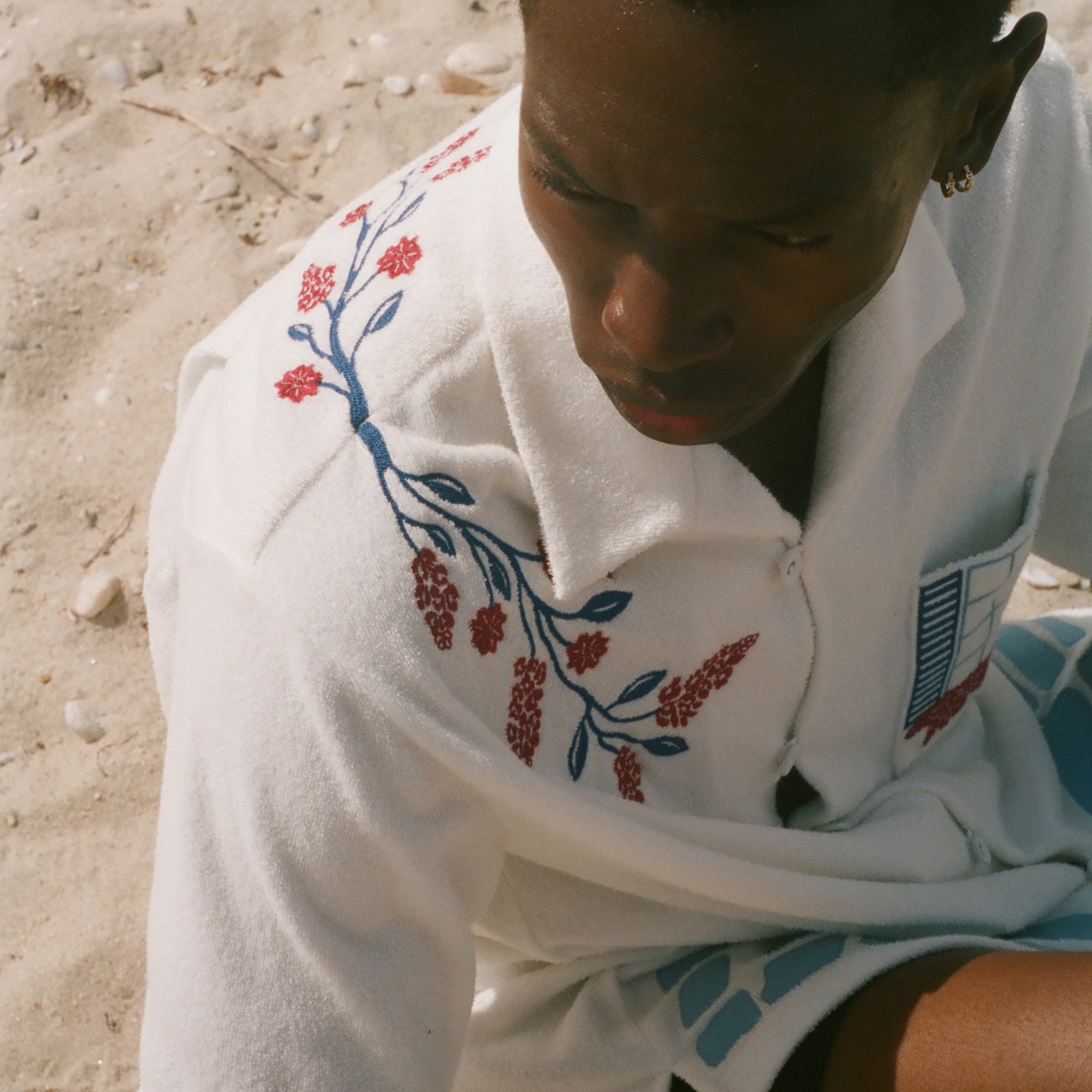 Overhead view of a person on a sandy beach wearing a cream terry cabana shirt with embroidered floral vine and window motifs.