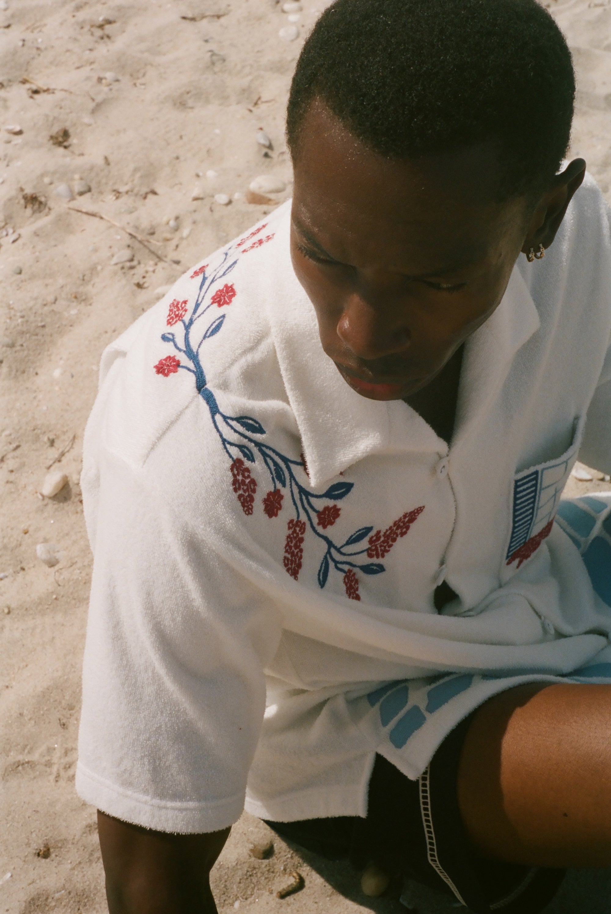 Overhead view of a person on a sandy beach wearing a cream terry cabana shirt with embroidered floral vine and window motifs.