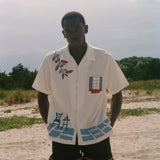 Person wearing a cream terry cabana shirt with embroidered floral, window, café, and cobblestone motifs on a sandy beach.
