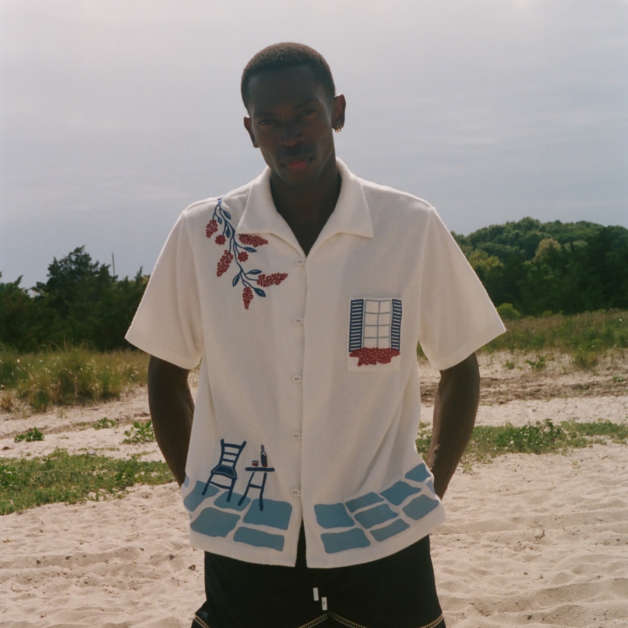 Person wearing a cream terry cabana shirt with embroidered floral, window, café, and cobblestone motifs on a sandy beach.