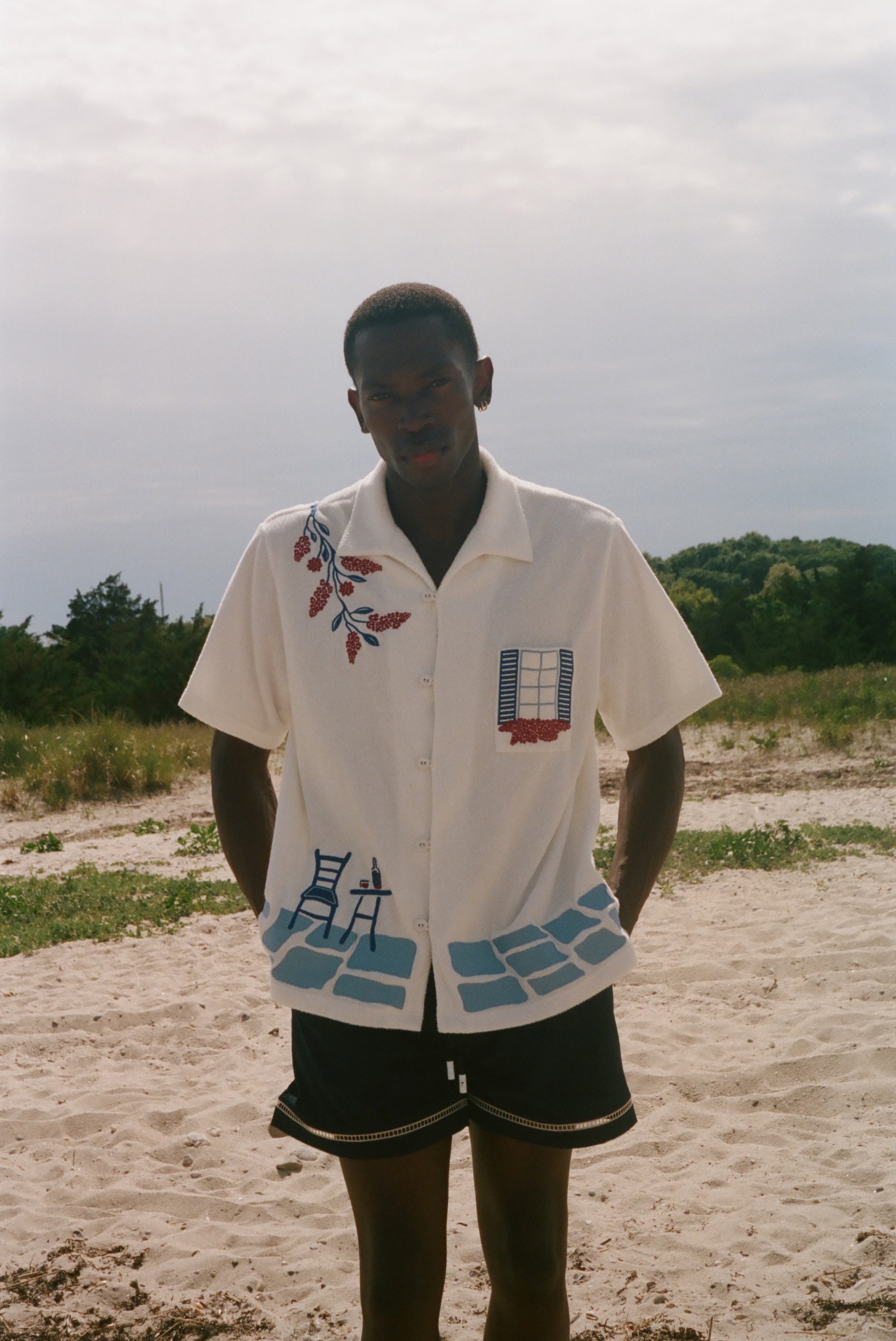 Person wearing a cream terry cabana shirt with embroidered floral, window, café, and cobblestone motifs on a sandy beach.