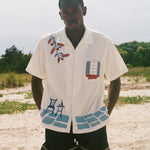 Man wearing a cream terry cabana shirt with embroidered floral, window, café, and cobblestone motifs on a sandy beach.