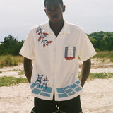 Man wearing a cream terry cabana shirt with embroidered floral, window, café, and cobblestone motifs on a sandy beach.