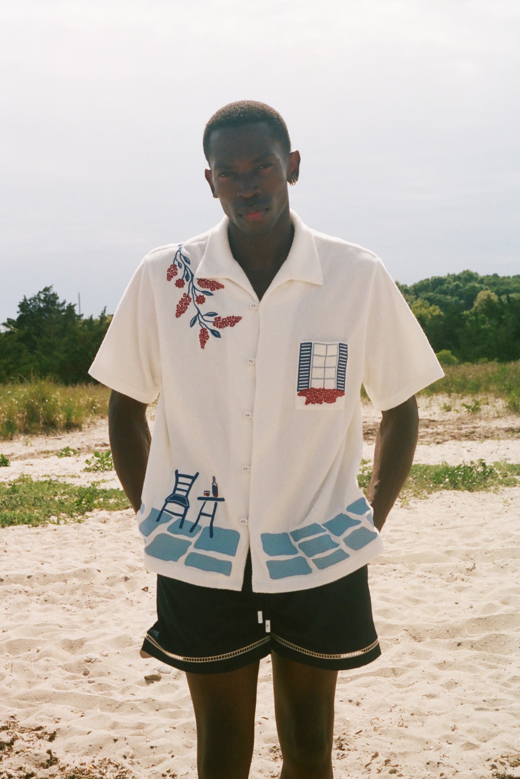 Man wearing a cream terry cabana shirt with embroidered floral, window, café, and cobblestone motifs on a sandy beach.