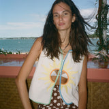A woman posing outdoors by the water wearing a sleeveless white vest with a yellow sun graphic and brown polka-dot pants.