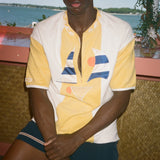 A man wearing a yellow and white color-blocked short-sleeve polo shirt with a geometric print, paired with dark shorts, seated in front of a scenic ocean background.