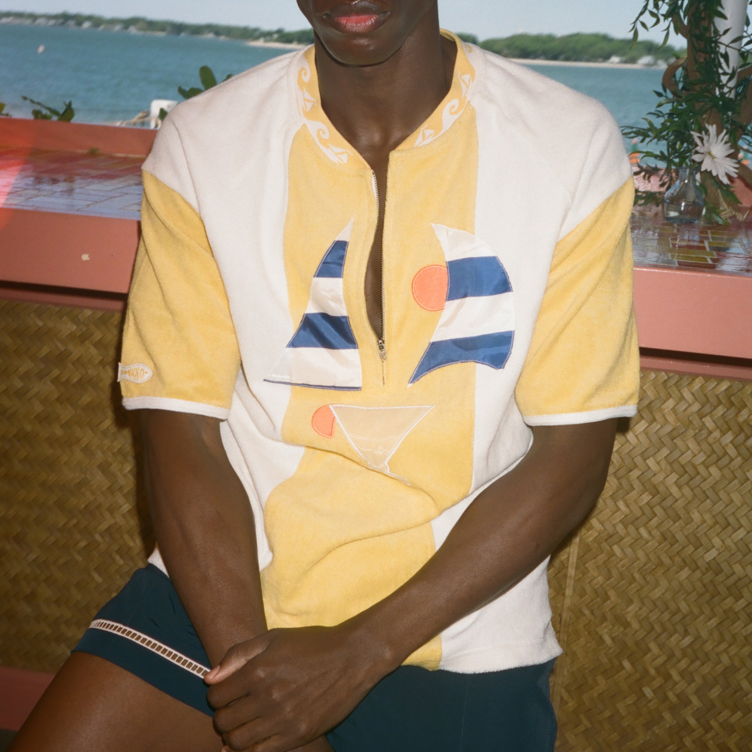 A man wearing a yellow and white color-blocked short-sleeve polo shirt with a geometric print, paired with dark shorts, seated in front of a scenic ocean background.