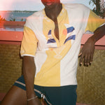 A man wearing a yellow and white color-blocked polo shirt with a cocktail graphic on the front, paired with dark teal shorts, posed against a tropical sunset background.