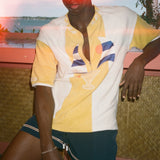 A man wearing a yellow and white color-blocked polo shirt with a cocktail graphic on the front, paired with dark teal shorts, posed against a tropical sunset background.