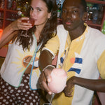 A man and a woman in a dimly lit bar setting, both wearing Sunset Beach collection items.