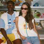 Two models posing together in a shop setting. The man wears a light blue and white striped polo shirt with yellow shorts, and the woman wears a white sleeveless top with a yellow sun graphic, denim shorts, and sunglasses.