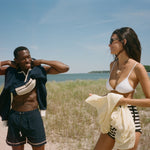 A man and a woman standing on a sandy beach dune with grass and the ocean in the background. The man is wearing navy blue swim trunks and has a navy blue shirt off his shoulders, while the woman wears a white bikini top and striped bottoms and holds a cream-colored shirt.