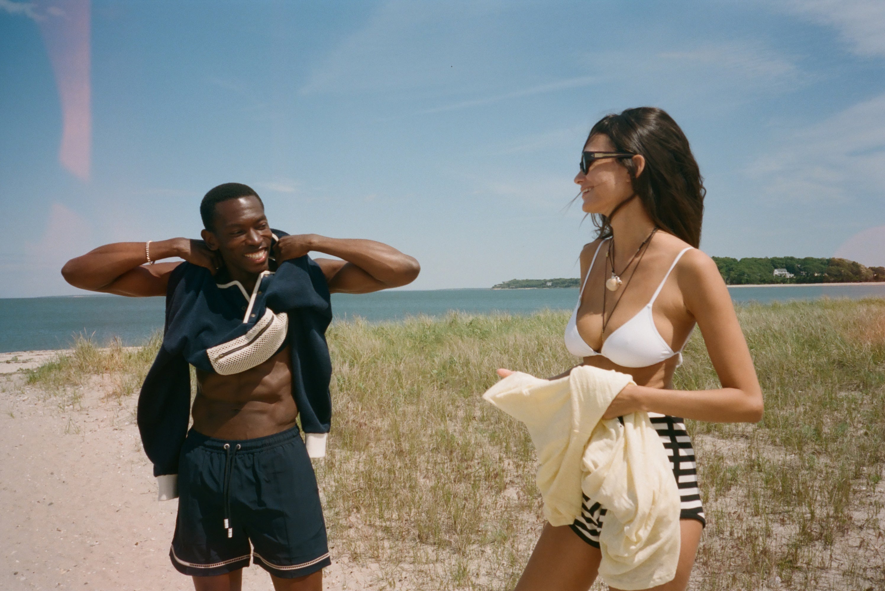 A man and a woman standing on a sandy beach dune with grass and the ocean in the background. The man is wearing navy blue swim trunks and has a navy blue shirt off his shoulders, while the woman wears a white bikini top and striped bottoms and holds a cream-colored shirt.