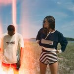 Two models standing outdoors in a grassy field under a blue sky. The model on the left wears a white t-shirt with 'TURISMO' printed on the chest and red shorts, while the model on the right wears a navy and white long-sleeve top with black and white striped shorts.