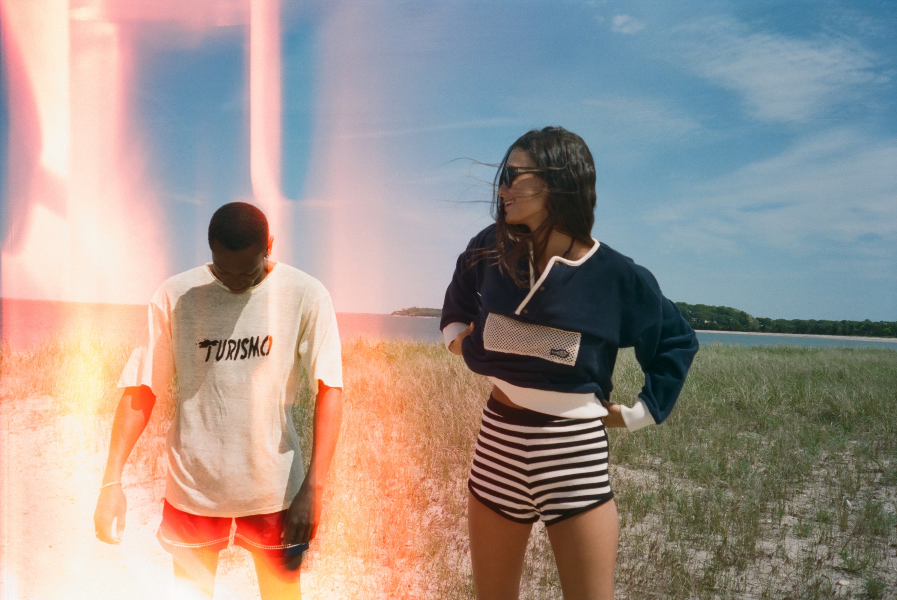 Two models standing outdoors in a grassy field under a blue sky. The model on the left wears a white t-shirt with 'TURISMO' printed on the chest and red shorts, while the model on the right wears a navy and white long-sleeve top with black and white striped shorts.