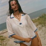 A woman stands on a sandy beach wearing a white short-sleeve button-front garment with blue trim along the collar, cuffs, and hem.