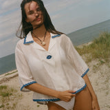 A woman stands on a sandy beach wearing a white short-sleeve button-front garment with blue trim along the collar, cuffs, and hem.